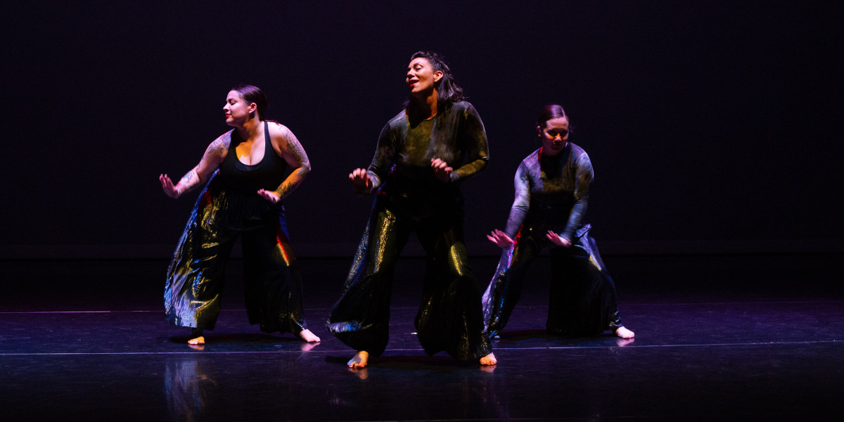 Three dancers on a darkly lit stage in a line, wearing silver pants dark shirts. They are standing with their knees and elbows bent, mid wiggle, with their hips jutting out to the side